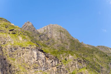 Pireneler 'deki açık mavi gökyüzünün altında sarp kayalık yamaçları ve yeşil bitki örtüsü olan sivri dağ sırtı. Güneş ışığı, dağlık arazinin engebeli dokularını ve keskin hatlarını vurgular.
