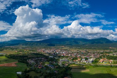 Laos 'un başkenti Vang Vieng' in geniş hava manzarası dramatik yeşil dağlarla dolu yemyeşil bir vadide kurulmuş ve canlı mavi gökyüzünün altında kümülüs bulutları yükselmekte. Sahne, kentsel gelişim ve çevre arasındaki zıtlığı vurguluyor