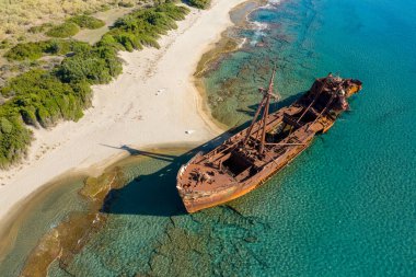 Yunanistan 'ın Mora kentindeki yemyeşil bitki örtüsü yakınlarındaki kumlu bir sahil boyunca sığ turkuaz sularda dinlenen paslı bir gemi enkazı ve kayalık deniz yatağı dokularının havadan görünüşü.