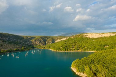 Bereketli ormanlık tepeler ve Bauduen yakınlarındaki dramatik kireçtaşı kayalıklarıyla çevrili turkuaz sularda yüzen yelkenlilerin bulunduğu huzurlu bir körfez manzarası..