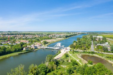 Geniş insansız hava aracı görüntüsü, Normandiya 'da berrak mavi gökyüzü altında yemyeşil, köy binaları ve açık kırsal alanlarla çevrili sakin bir nehri çevreleyen Pegasus Köprüsü' nü gösteriyor..