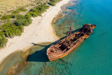 Dimitrios 'un gemi enkazının yüksek açılı görüntüsü kristal berraklığında turkuaz sularda Yunanistan' ın Gythio kenti yakınlarındaki el değmemiş kumlu bir sahil ve yemyeşil bitki örtüsünün yanında. Paslı dokular ve canlı kıyı renkleri parlak gün ışığıyla aydınlatılır..