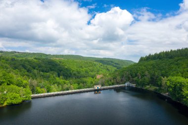 Beton barajın panoramik görüntüsü, yoğun yeşil ormanlık ve Chaumecon gölünde dramatik bulutlarla dolu gökyüzünün altındaki yuvarlanan tepelerle çevrili, derin ve durgun bir göl boyunca uzanır..