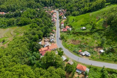 Curving country road winds through a small village with red-roofed houses, surrounded by lush green hills and dense forest in northern Laos. Terraced rice fields and scattered homes highlight the tranquil rural landscape under bright daylight.
