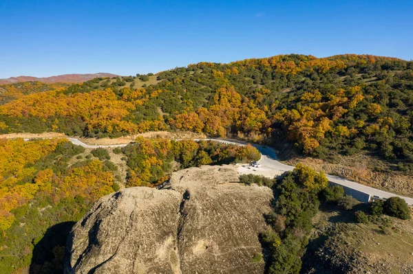 Yunanistan 'ın anakarasının güneşli bir yamacında, engebeli bir kaya oluşumunu ve canlı bir sonbahar ormanını aşan, açık mavi gökyüzü altında kıvrımlı bir yolun hava görüntüsü..