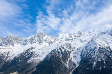 Sharp, rugged summits of the Mont Blanc massif rise above a glacier and dark forested slopes, with textured clouds drifting across a bright blue sky.