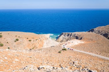 Girit 'teki Seitan Limania plajındaki canlı mavi Akdeniz' e doğru uzanan kurak ve kayalık arazide virajlı bir yol ve açık bir gökyüzünün altında parkedilmiş arabalar görülüyor..