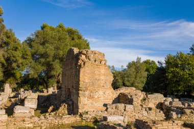 Eski harabelerden kalma tuğla ve taş duvarlar Yunanistan 'ın Olympia kentindeki dağınık taş ve yemyeşil duvarlar arasında yer alıyor. Sıcak öğleden sonra güneşi, bu sakin arkeolojik manzaradaki dokulu yüzeyleri ve olgun ağaçları vurguluyor..