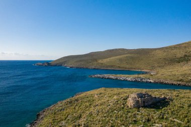 Açık mavi sular Yunanistan 'ın Cap Tenare kentinde engebeli tepeler ve kayalık kıyı şeridi ile buluşuyor. Güneş ışığı, uçsuz bucaksız bulutsuz gökyüzünün altında vahşi otları ve ıssız bir taşı vurgular..