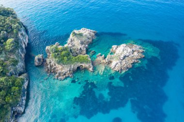 Yunanistan 'ın Epirus kentindeki Karavostasi kıyı şeridi boyunca engebeli kayalık adaları ve yemyeşil bitki örtüsünü berrak turkuaz su kaplar. Parlak gün ışığı, canlı Akdeniz sahili ve doğal kıyı şeridini vurguluyor.