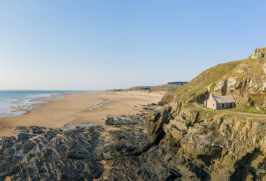 Cap de Carteret 'teki kayalık kayalıkların tepesinde, engin boş bir sahile ve berrak bir gökyüzünün altında sakin bir mavi denize bakan yalnız bir taş ev bulunur. Sahne, engebeli dokuları ve açık, sakin kıyı şeridini vurgulayan hafif gün ışığında yıkanır..