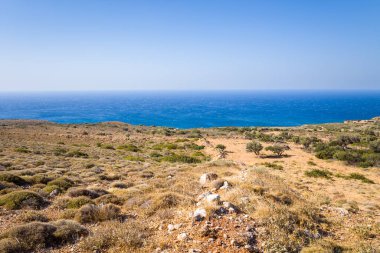 Sunlit arid terrain with scattered shrubs and stones stretches toward the deep blue Mediterranean Sea under a clear sky on the southern coast of Crete. The scenery is open and windswept, evoking a sense of wild solitude.