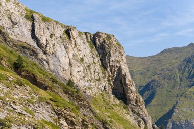 Güneş ışığıyla aydınlatılan dik kayalık uçurum, Pireneler 'deki Gavarnie Gedre yakınlarındaki yeşil dağlık yamaçlardan yükseliyor. Sahnede desenli taş, seyrek bitki örtüsü ve açık mavi bir gökyüzü yer alıyor..