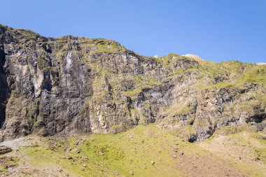 Pirene Dağları 'ndaki yeşil eğimli bir tepenin üzerinde yükselen engebeli bir kayanın geniş manzarası açık gün ışığı ve parlak mavi bir gökyüzü tarafından vurgulanıyor. Çimenlerin ve taşların zıt dokuları çarpıcı bir dağ manzarası yaratır..