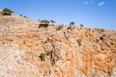 Girit 'teki Seitan Limania' daki engebeli turuncu kireçtaşı kayalığı, açık mavi gökyüzünün altında küçük çalılar ve ağaçlarla süslenmiş. Dokulu kaya yüzeyi ve en az yeşillik kurak bir Akdeniz manzarasını çağrıştırıyor.