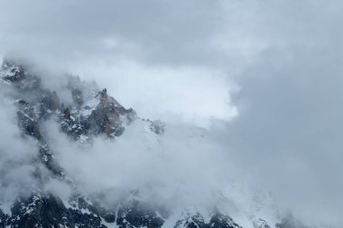 Aiguille du Midi 'nin keskin, karla kaplı kaya kuleleri yoğun sis ve girdap bulutları tarafından kısmen gizlenerek gizemli ve sakin bir alp atmosferi oluşturur..