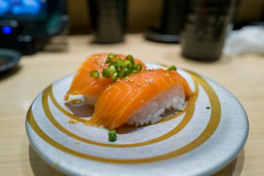 İki parça somon nigiri sushi, üzerinde doğranmış yeşil soğan ve susam tohumları modern gümüş ve altın tepside servis edilir. Parlak balıklar ve narin pirinçler çağdaş bir restoranda hafif kapalı mekan ışıklarıyla vurgulanır..