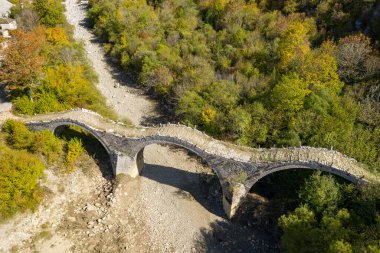 Yunanistan 'ın Epirus kentindeki Kipoi yakınlarında sık sonbahar yapraklarıyla çevrili kuru, kayalık bir nehir yatağını geçen tarihi üçlü kemerli Plakidas Köprüsü' nün yüksek perspektifi. Parlak gün ışığı, dokulu taş işçiliğini ve canlı manzarayı vurgular.