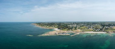 Saint-Gildas-de-Rhuys 'taki Port aux Moines plajı ve marinasının geniş insansız hava aracı manzarası, berrak turkuaz su, kayalık kıyı şeridi ve parlak bir gökyüzü altında genişleyen bir kıyı köyü bulunuyor..