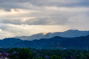 Yoğun bulut örtüsü ve yumuşak akşam ışığı Vang Vieng, Laos 'ta katmanlı, ormanlı tepeler ve dağınık çatılar üzerinde dramatik bir atmosfer yaratır. İnce ışınlar yemyeşil manzarayı ve uzak tepeleri aydınlatır..