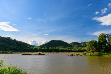 Çamurlu Mekong nehrinin geniş bir bölümü Laos 'un Luang Prabang yakınlarındaki yeşil tepeleri ve yoğun nehir kenarı bitkilerini geçerek akar. Geleneksel bir longboat canlı mavi gökyüzünün altında sürükleniyor, dağınık bulutlarla, canlı bir doğal manzara yaratıyor..