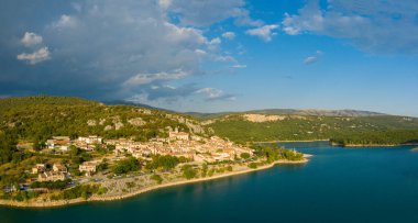 Bauduen köyünün hava manzarası sıcak akşam güneşiyle aydınlanıyor canlı turkuvaz suları ve parçalı bulutlu gökyüzünün altındaki yemyeşil tepelerle..