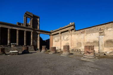 Antik tuğla ve taş sütun temelleri Pompeii, İtalya 'nın açık bazilika avlusunda duruyor. Parlak güneş ışığı, klasik Roma mimarisini, dokulu duvarları ve yıpranmış mavi gökyüzünü vurgular..