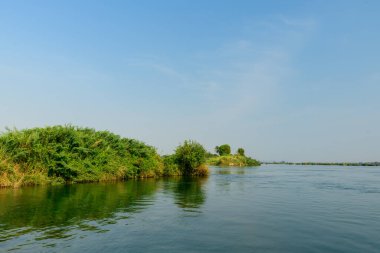 Lush green islets rise from the calm waters of the Mekong River in the 4000 Islands region of southern Laos. Clear daylight and a vast blue sky create an open, tranquil riverscape with natural reflections and vibrant vegetation.