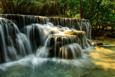 Yumuşak güneş ışığı kuzey Laos 'taki Kuang Si Şelaleleri' ndeki kireçtaşı teraslarını ve temiz havuzu vurguluyor. Bereketli tropikal orman çağlayan suyu çerçeveler ve katmanlı dokularla sakin bir doğal manzara yaratır..