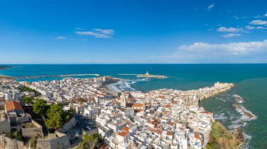 İtalya 'nın Vieste kentinin geniş panoramik manzarası, turkuaz Adriyatik Denizi üzerindeki kireçtaşı kayalıklarda duran ve Santa Eufemia deniz fenerinin küçük bir açık deniz adasında durduğunu ortaya koyuyor. Sahnede katmanlı çatılar var.