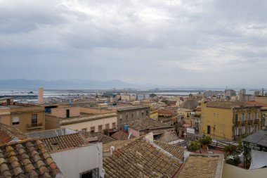 Cagliaris 'in eski çatılarına geniş bir perspektif. Döşeme kiremitleri var. Yoğun limana ve bulutlu gökyüzünün altındaki uzak dağlara bakıyor..