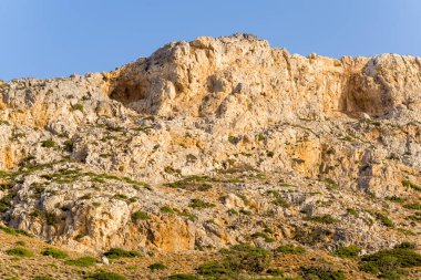Sıcak güneş ışığı Girit, Balos yakınlarındaki dağlarda kayalık bir kayalık yüzeyin engebeli dokularını ve toprak tonlarını vurgular. Sade bitki örtüsü, berrak mavi bir gökyüzünün altında sivri kireçtaşı yüzeyine saçılmıştır..