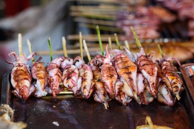 Bambu çubuklarıyla şişlenmiş taze ızgara mürekkep balığı, Kep, Kamboçya 'daki hareketli bir deniz ürünleri standında metal bir tepside düzenli sırada diziliyor. Görüntü kömürleşmiş dokuları, parlak yüzeyleri ve Güneydoğu Asya caddesinin canlı atmosferini vurguluyor