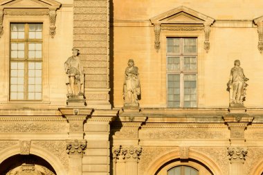 Fransa 'nın Paris kentindeki Louvre Müzesi' nin tarihi cephesinde süslü kemerli camların üzerine üç adet klasik taş heykel yerleştirilmiştir. Ilık altın güneş ışığı, karmaşık oymalar, dekoratif sütunlar ve desenli duvarları vurguluyor.
