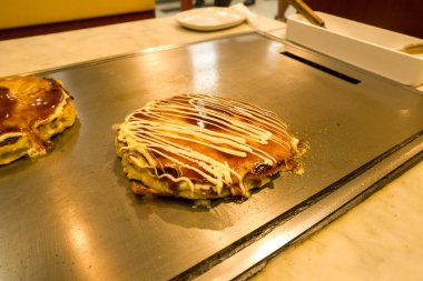 Japanese okonomiyaki pancake topped with savory sauce and mayonnaise is cooked on a hot metal griddle in a restaurant setting. Warm lighting highlights the golden, crispy edges and glossy surface of the dish.