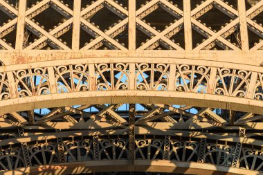 Eiffel Kulesi 'ndeki süslü demir kemerlerin ve kafes işçiliğinin detaylı görüntüsü, sıcak günbatımı ışığı ile aydınlatılıyor ve yıpranmış metal yüzey boyunca karmaşık gölgeler bırakıyor..