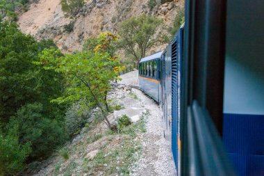 Yunanistan 'ın Kalavrita kenti yakınlarındaki yeşil ağaçlar ve engebeli kayalık yamaçlarla çevrili çakıllı tren yolu boyunca mavi dar ölçülü bir tren kıvrımları bulunuyor. Sahne, yemyeşil yapraklarla Mora dağlarının dokulu kayalıkları arasındaki zıtlığı vurguluyor..