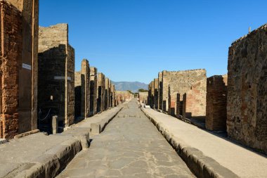 İtalya 'nın Pompeii kentindeki arkeolojik kamaralarda yıpranmış tuğla ve volkanik taş duvarlarla kaplı Roma taşlı caddeye uzun bir perspektif. Parlak güneş ışığı ve açık mavi gökyüzü geometrik çizgileri ve tarihi dokuları gözler önüne seriyor.