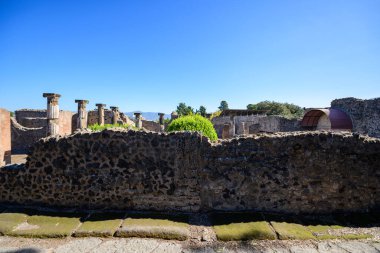 Yıpranmış volkanik taş duvarlar ve klasik sütunlar Pompeii arkeolojik sahasında canlı bir mavi gökyüzünün altında duruyor. Sahnede güneş ışığı alan duvarcılık, dağınık yeşillik ve tarihi Roma harabelerinin açık manzarası yer alıyor..