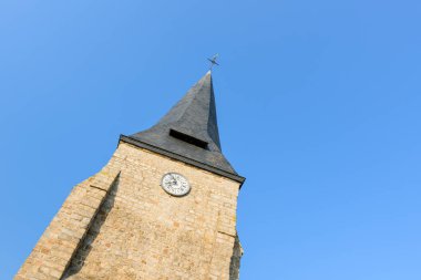 Saint Sylvain 'da parlak mavi gökyüzüne karşı kurulu tarihi bir taş kilise kulesinin, tahta kule ve saat yüzlü görüntüsü. Sıcak güneş ışığı, yapılandırılmış duvarcılığı vurguluyor. Huzurlu ve zamansız bir atmosfer yaratıyor..