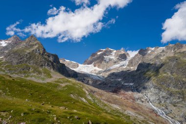 Buzul de la Lee Blanche ve dramatik Aiguilles de Tre la Tete 'yi, yemyeşil çayırlar, kayalık tepeler ve açık mavi gökyüzünün altında çarpıcı bir buzul, dağınık beyaz bulutlar..