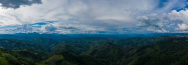 Sweeping panorama of densely forested hills and distant blue ridges beneath a dramatic sky filled with layered clouds in the rural region between Phonsavan and Phou Khoun, Laos. The scene highlights natural contours, lush vegetation, and the vastness
