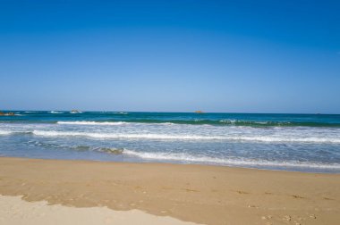 Yumuşak dalgalar Kumihamacho 'daki Shotenkyo Sahili' nin temiz kumlu sahillerinde dalgalanır, üzerinde canlı mavi bir gökyüzü uzanır ve ufukta küçük kayalık adalar görünür. Sahne parlak, açık ve davetkar..