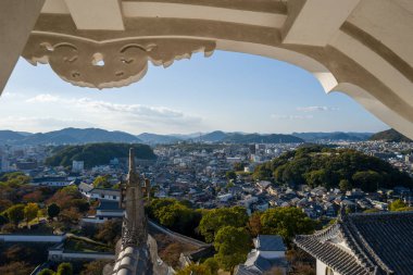 Himeji Kalesi 'nin dekoratif beyaz saçakları şehrin panoramik manzarasının üstünde, kiremitli çatıları, yeşil tepeleri ve berrak gökyüzünün altındaki uzak dağları var..