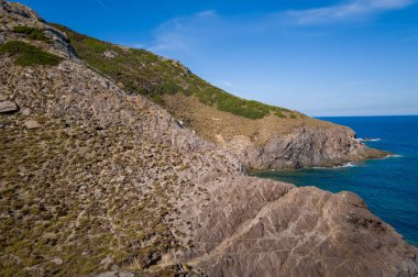 Ender bitki örtüsüyle açılı kayalık burunlar Arjantin, Sardunya yakınlarındaki canlı mavi Akdeniz ile kesişiyor. Sahnede engebeli dokular, güneşli yamaçlar ve berrak bir gökyüzünün altında sakin bir kıyı atmosferi yer alıyor..