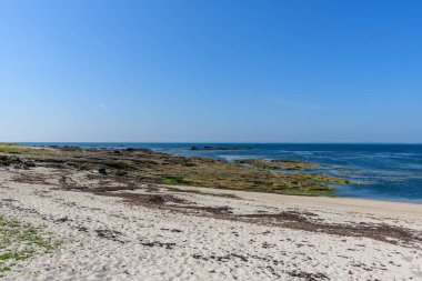 Wide sandy beach meets a rugged rocky shoreline at Plage de la Jument in Quiberon, with clear blue water and a cloudless sky creating a tranquil coastal scene.