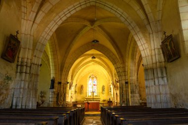 Sıcak ışık, Gotik kemerleri, ahşap sıraları ve sunağın üstündeki renkli camlı pencereleri vurgulayan tarihi bir taş kilisenin kapağını dolduruyor. Yıpranmış duvarlar ve dini heykeller sessiz bir miras ve zamansız bir anlayış yaratır.