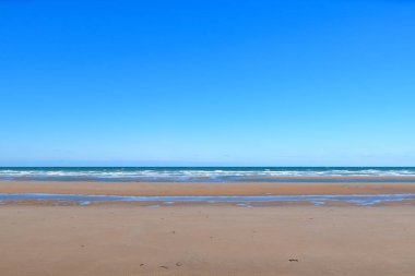 Geniş kumlu Omaha Sahili ufuk çizgisine kadar uzanır, hafif dalgalar dalgaların dalgalı mavi gökyüzünün altında kıyıya vurmasıyla. Sahne açık, sakin ve dağınık, bir boşluk ve sükunet hissi uyandırıyor..