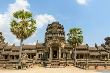 Angkor Wat 'ın ana kumtaşı giriş pavyonu iki uzun palmiye ağacı arasında simetrik olarak duruyor parlak mavi gökyüzü altında dağınık bulutlar. Süslü oymalar, yıpranmış taşlar ve geometrik çizgiler bu ikonik Khmer 'in ihtişamını vurgular.