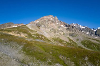 A rugged mountain ridge rises above rolling green and rocky slopes at Col de la Seigne, under a deep blue sky. The bright sunlight emphasizes the contrasting textures of grass, stone, and patches of lingering snow.